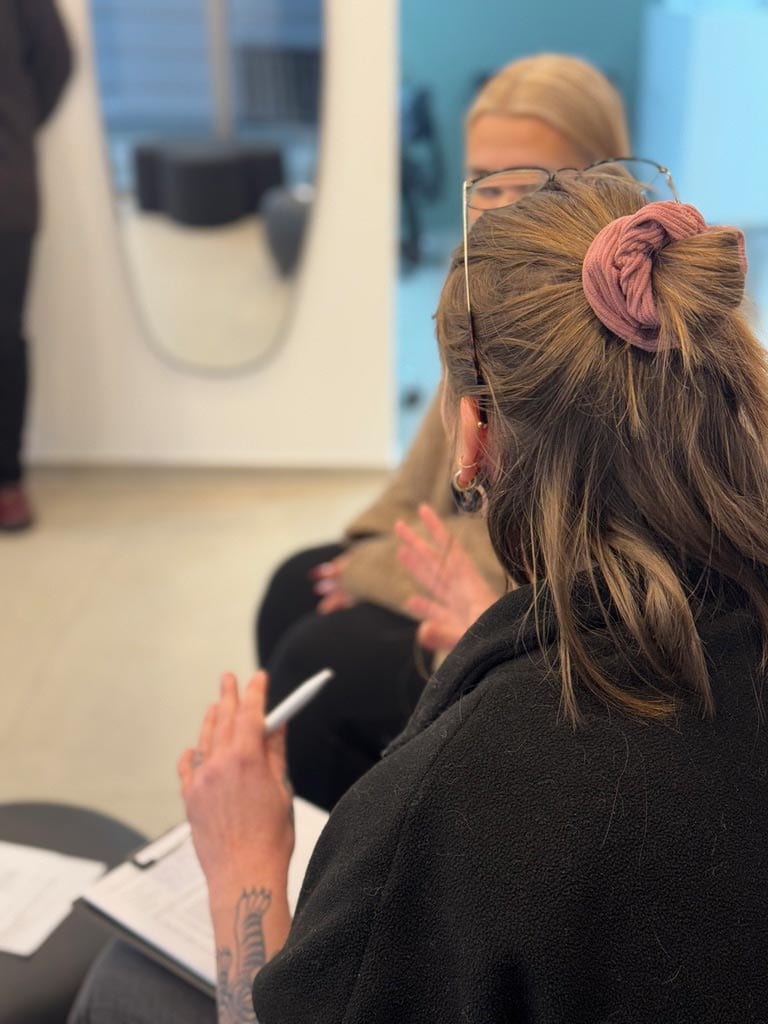Person mit Tattoo und rosa Haargummi spricht, hält einen Stift, unscharfer Hintergrund.