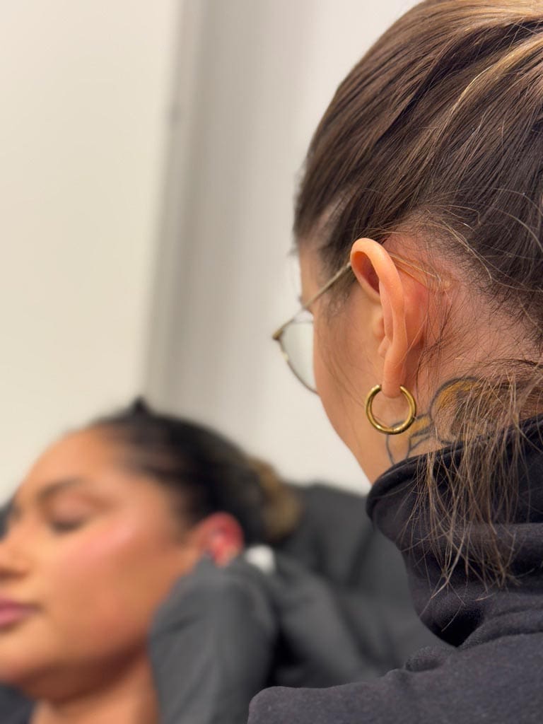 Zwei Frauen, eine mit Brille; eine lächelnd im Profil. Goldene Ohrringe, unscharfer Hintergrund.