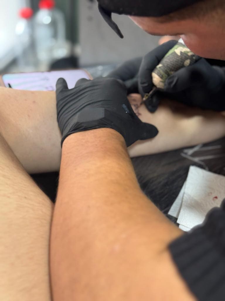 Person tätowiert einen Arm, trägt schwarze Handschuhe; Haut hell. Konzentration sichtbar.